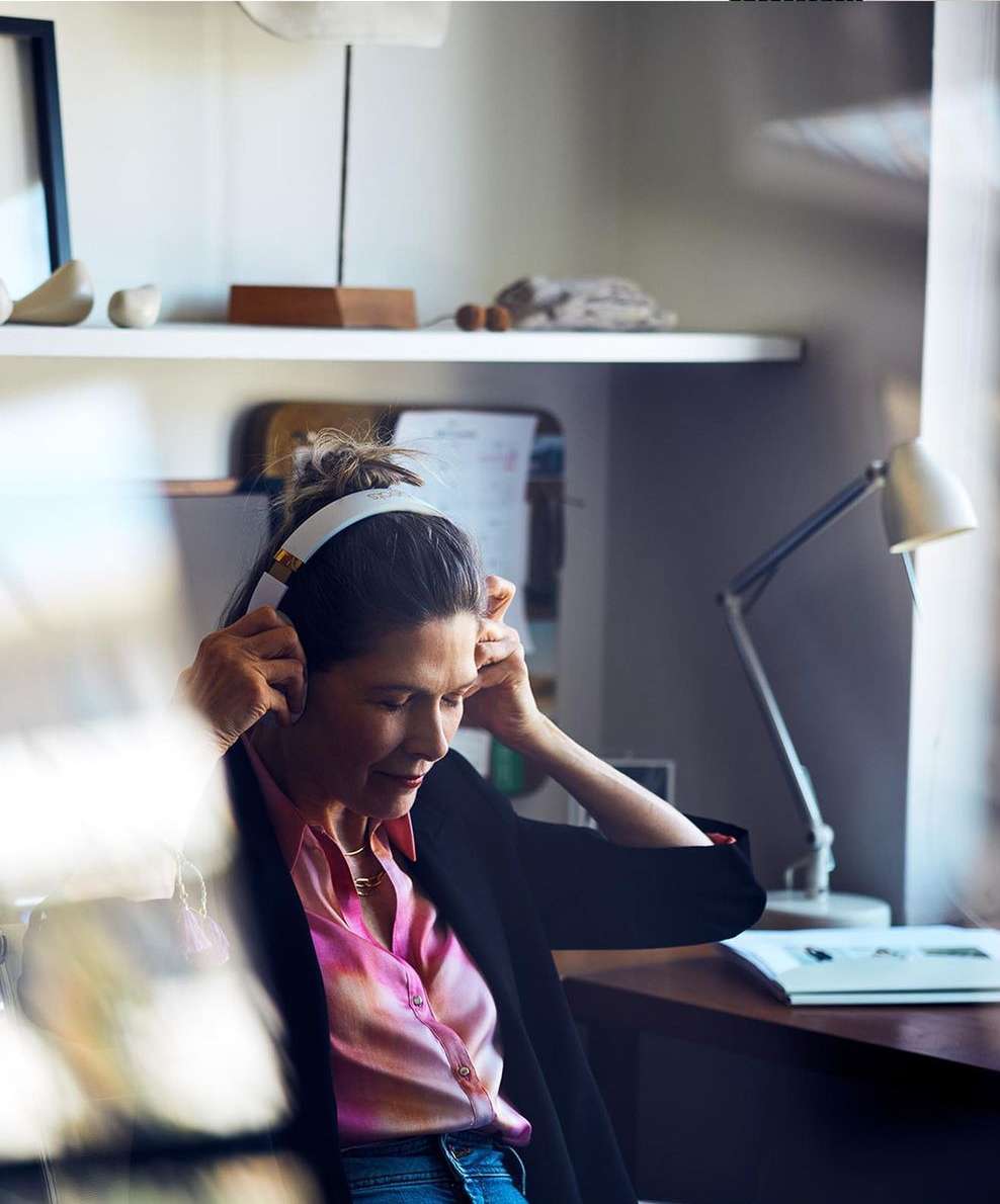Woman with headphones sitting at a desk with a lamp, artwork on shelves, wearing a pink shirt and blazer in an indoor setting.