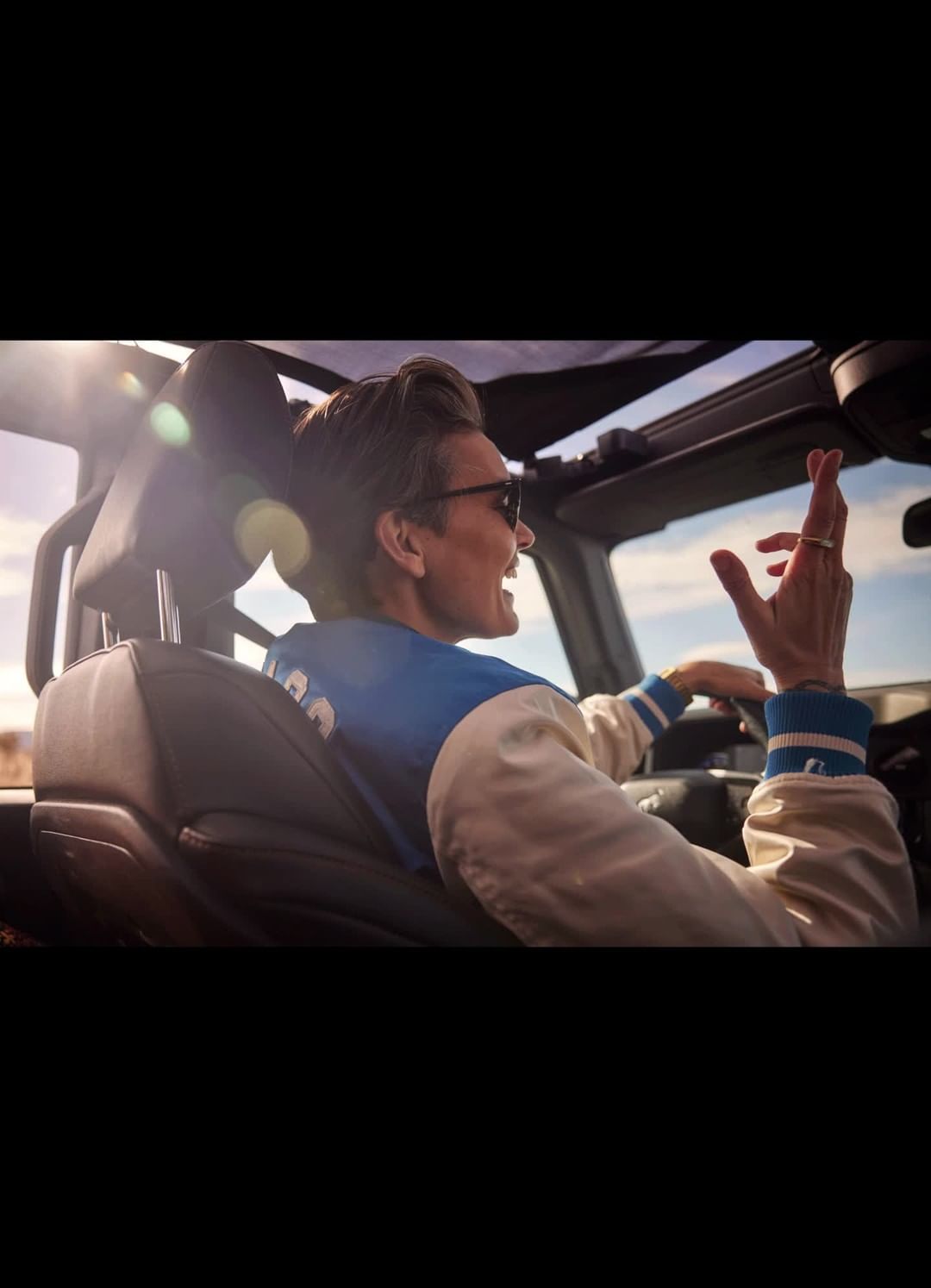 A man driving a convertible car on a sunny day, wearing a bomber jacket and sunglasses, with a landscape visible in the background.