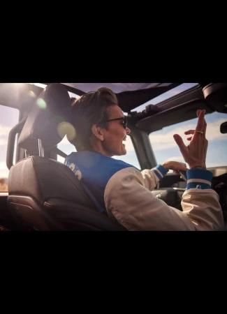 A man driving a convertible car on a sunny day, wearing a bomber jacket and sunglasses, with a landscape visible in the background.