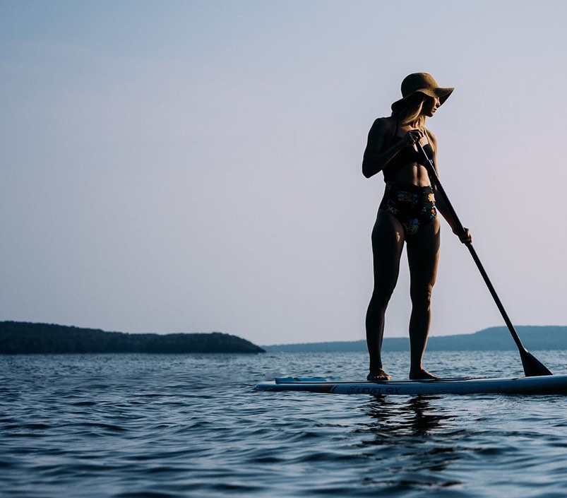 A woman in a hat stands on a paddleboard with a paddle in hand, silhouetted against a bright sky and water landscape.
