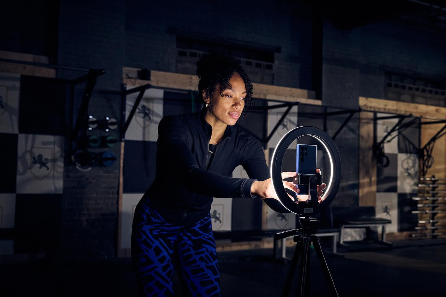A woman uses a ring light and her smartphone for content creation, set against an industrial gym backdrop, for fitness or technology.