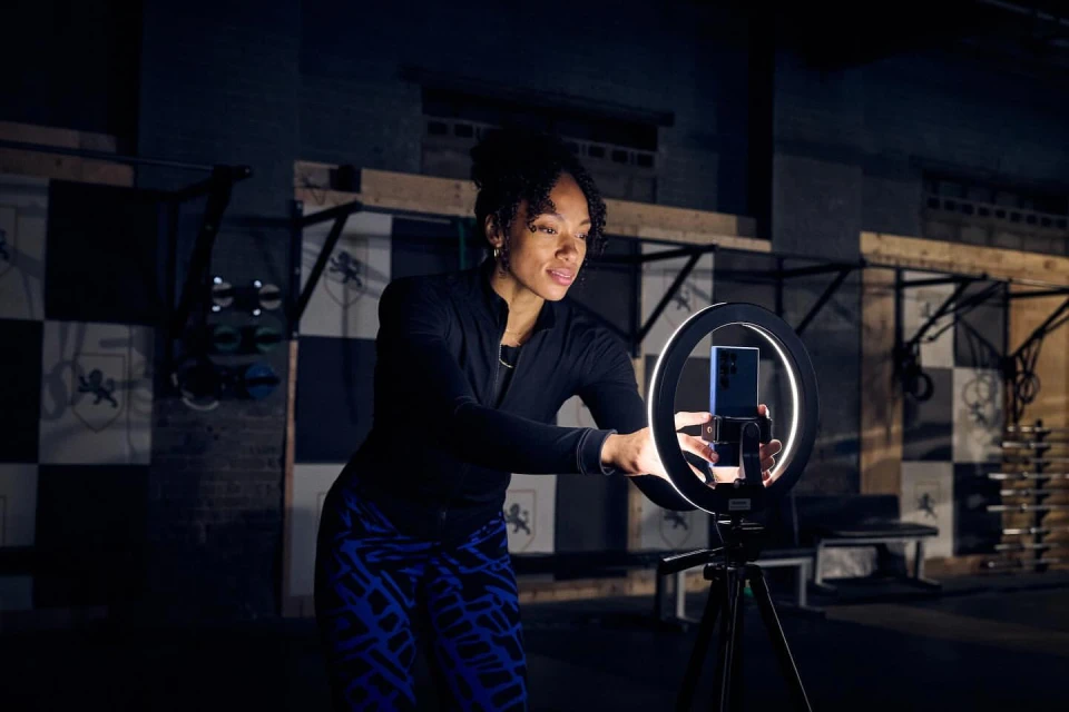 A woman uses a ring light and her smartphone for content creation, set against an industrial gym backdrop, for fitness or technology.
