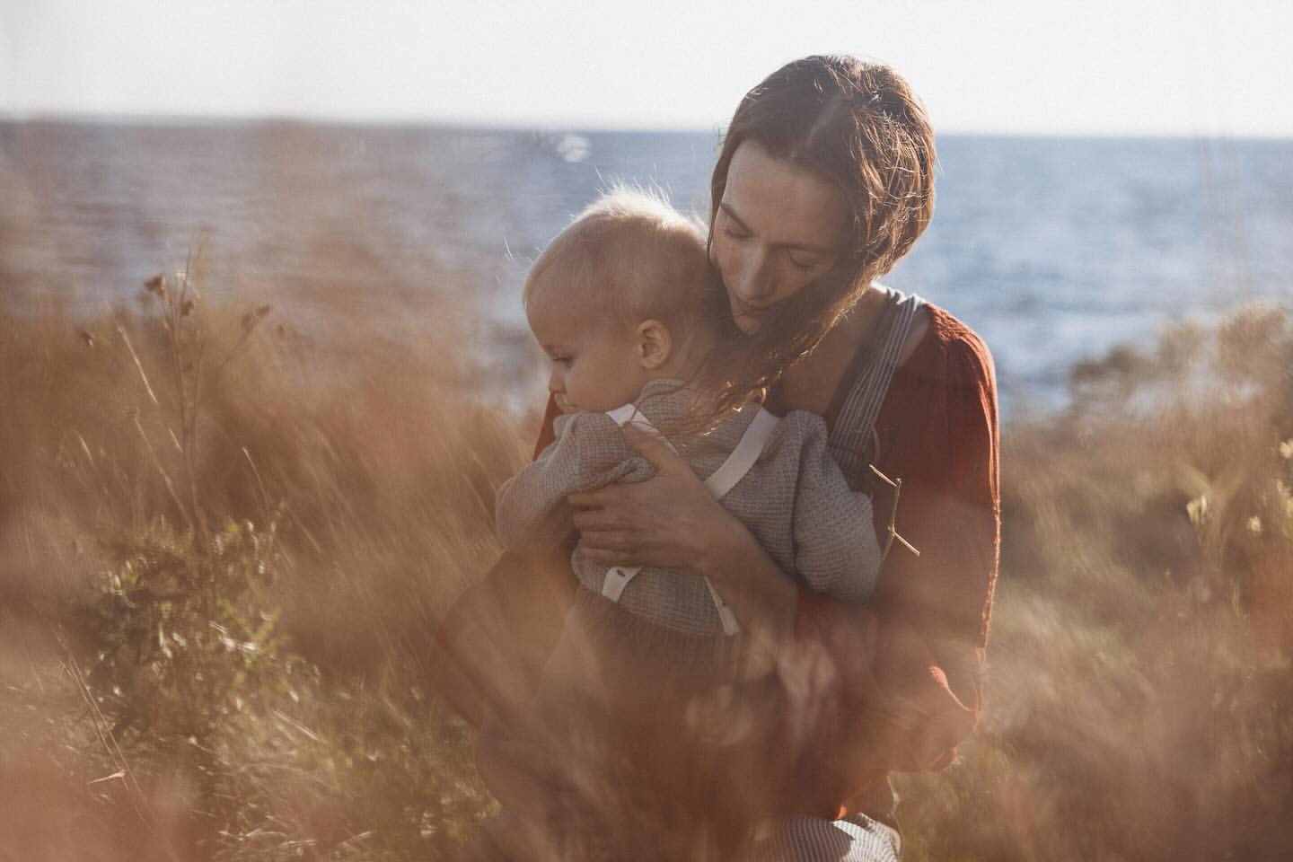 A mother holds her baby outdoors in soft light, standing in a field of tall brown grass with a blurred ocean backdrop, showcasing a tender, natural scene.