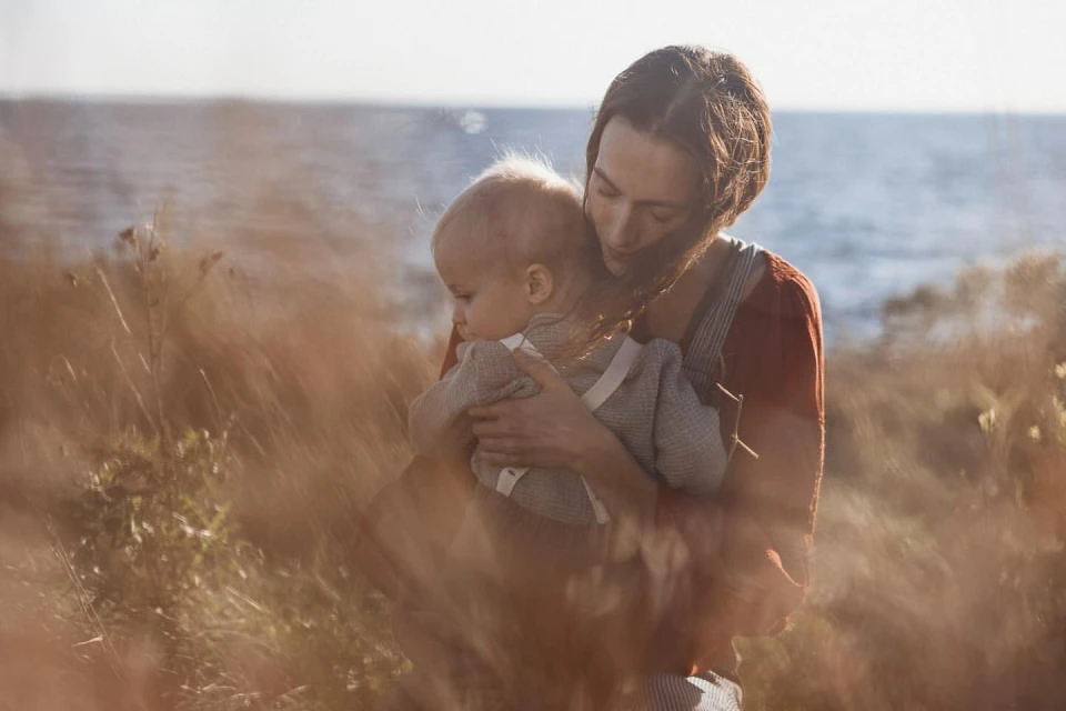 A mother holds her baby outdoors in soft light, standing in a field of tall brown grass with a blurred ocean backdrop, showcasing a tender, natural scene.