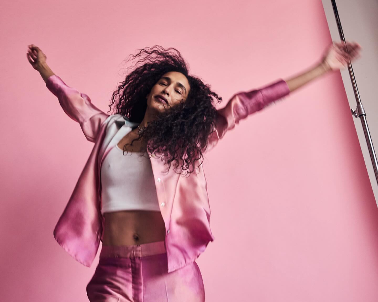 A woman with curly hair in a pink satin suit is dancing with her arms raised against a pink background in a studio environment.