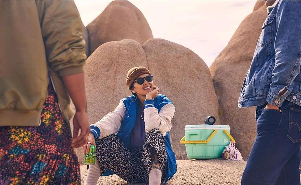 Woman in beanie sitting among rock formations with a drink, a cooler, and two partially-visible standing people in outdoor setting.