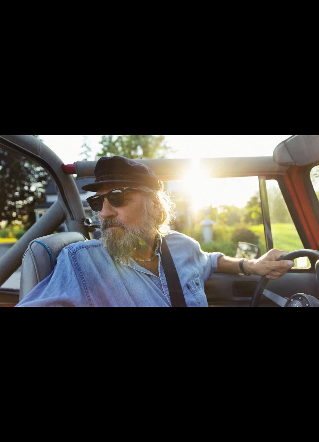 An older adult male drives a convertible on a sunny day, wearing sunglasses and a cap, with green trees and a house in the background.