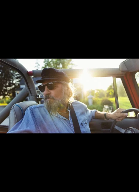 An older adult male drives a convertible on a sunny day, wearing sunglasses and a cap, with green trees and a house in the background.