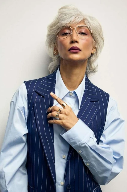 Portrait of stylish older woman in pinstripe vest and eyeglasses, confidently posing against a clean, neutral backdrop.