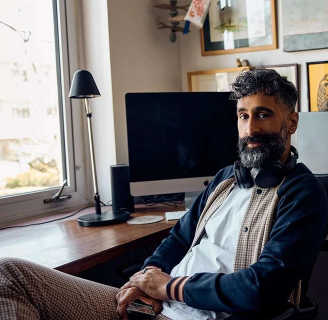 Man with beard sits at desk with computer, lamp, and wall art, wearing headphones and varsity jacket in indoor setting.