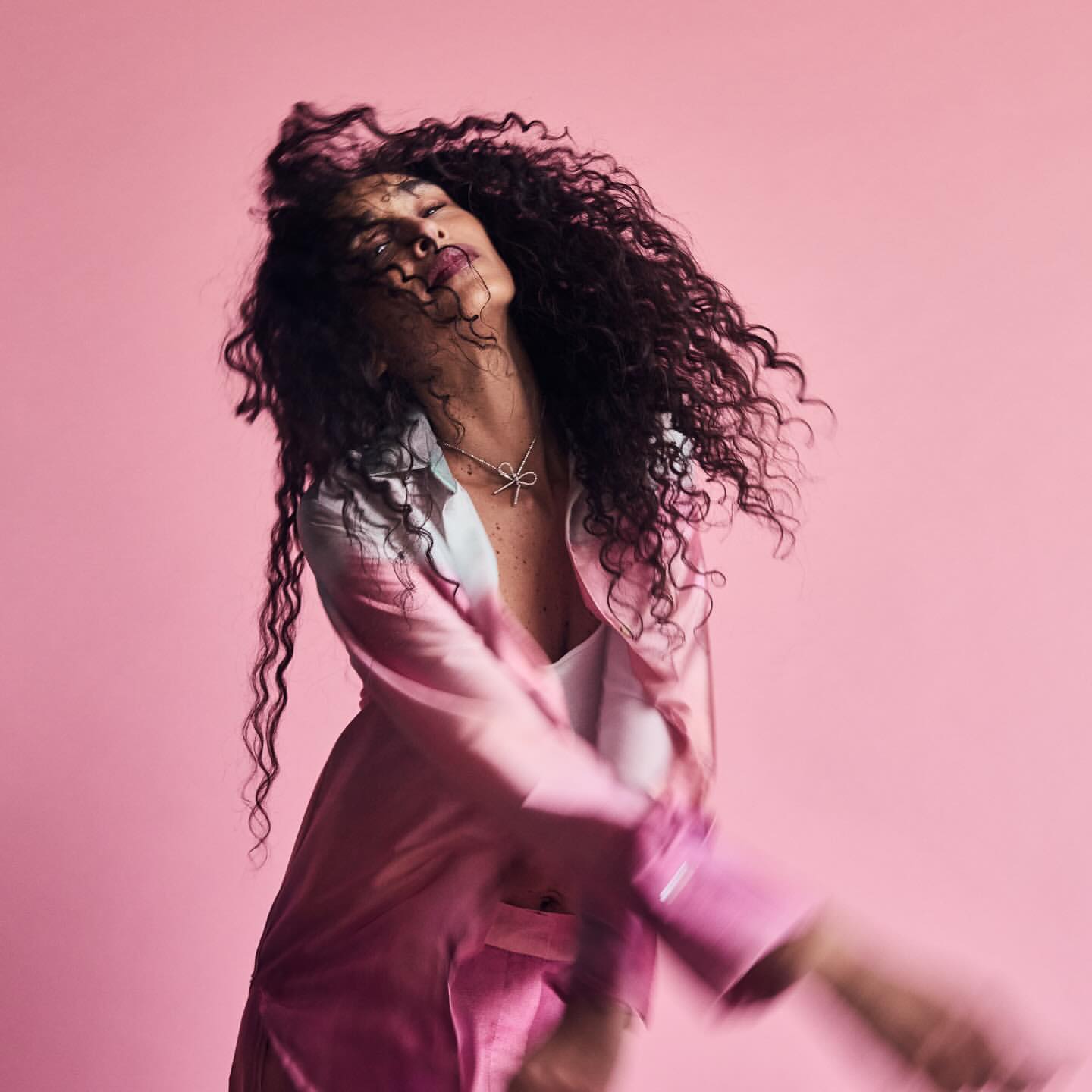Woman with curly hair in motion, wearing pink top and pants against a pink background, accessorized with a bow necklace and pink lipstick.