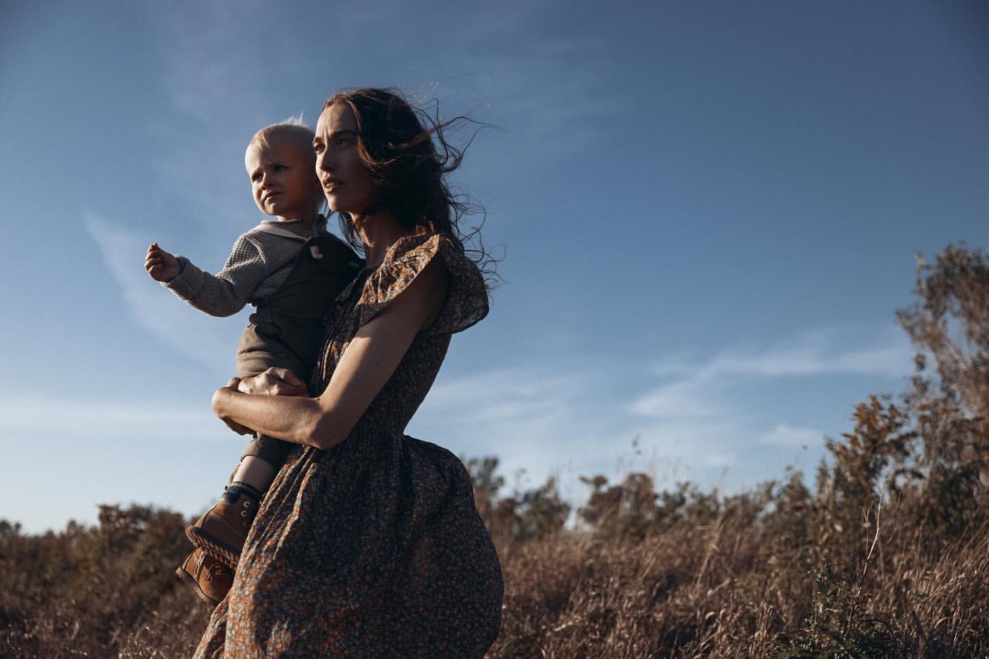 A woman in a dress holds a young child in an outdoor field under a bright blue sky, with golden light enhancing the scene's warmth.