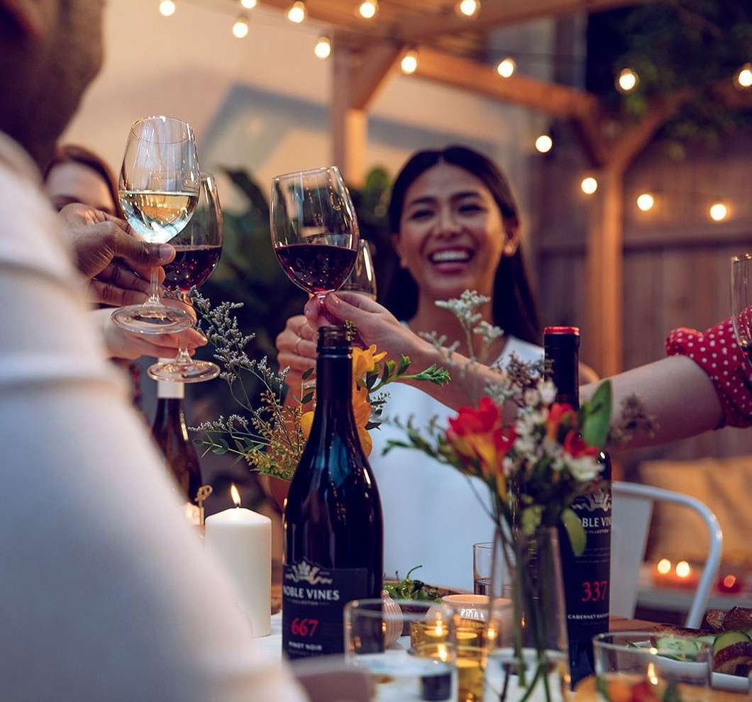 People toasting with wine glasses at an outdoor gathering. Festive atmosphere with string lights and wine bottles on a table.