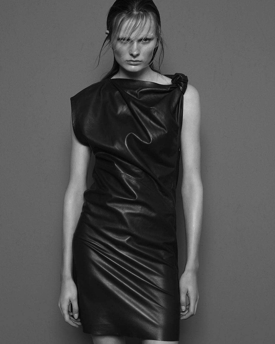 Black and white studio portrait of an adult female model wearing a black leather dress, standing against a solid grey background.