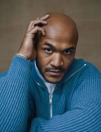 Portrait of a bald Black man wearing a blue ribbed zip-up sweater with his hand resting on his head.