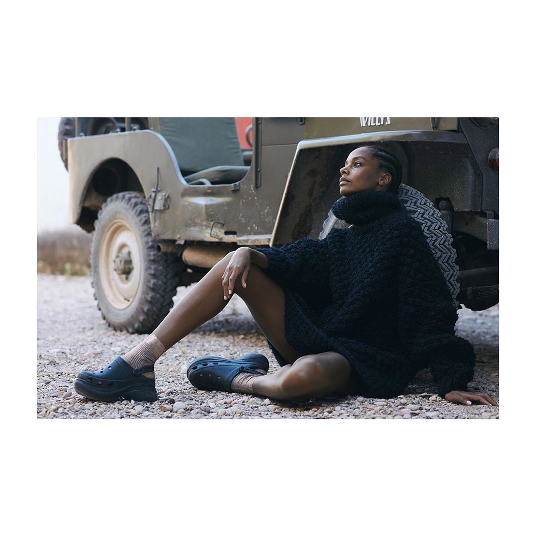 Adult female model posing in a black knit dress and black shoes sitting next to a vintage military jeep on a gravel surface.