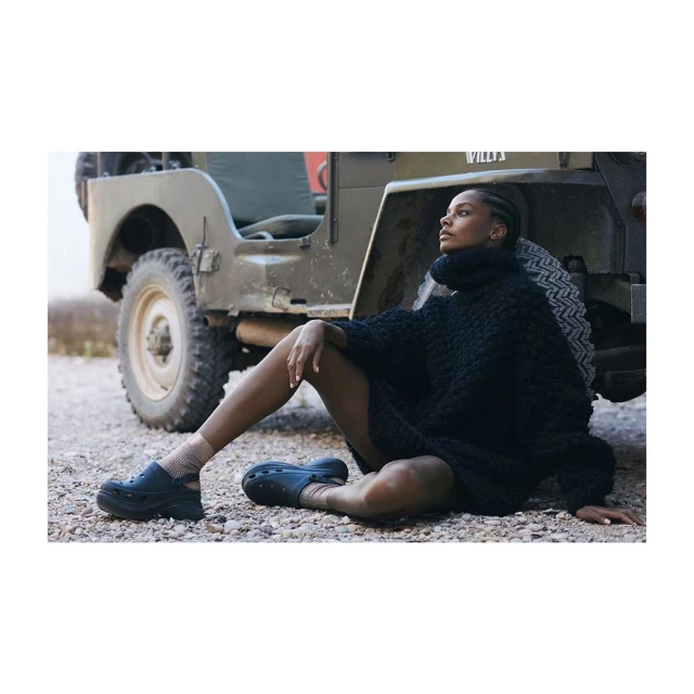 Adult female model posing in a black knit dress and black shoes sitting next to a vintage military jeep on a gravel surface.