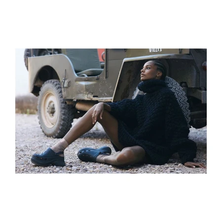 Adult female model posing in a black knit dress and black shoes sitting next to a vintage military jeep on a gravel surface.