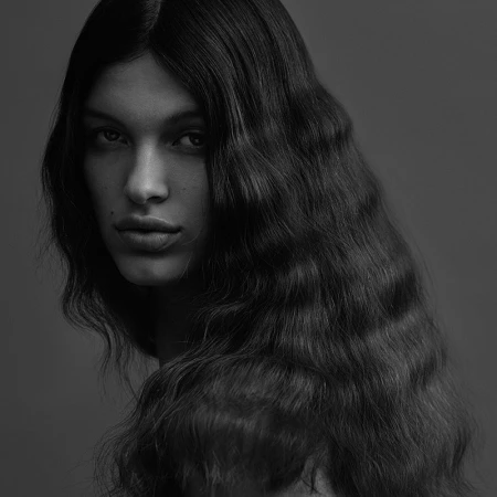 Portrait of a woman with long dark wavy hair looking over her shoulder against a plain grey background
