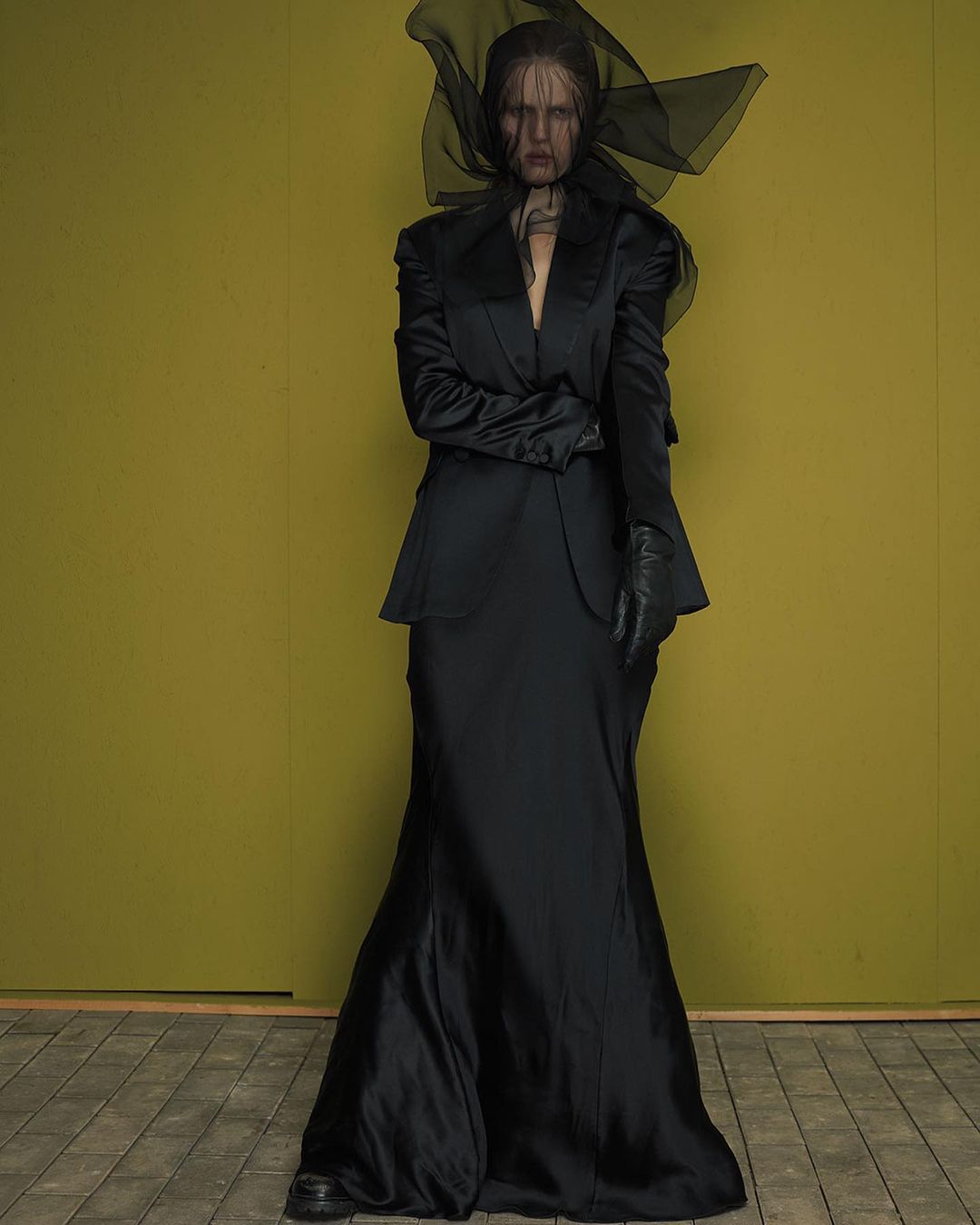 Model in black satin suit and sheer veil headpiece standing against an olive backdrop