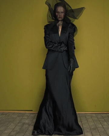 Model in black satin suit and sheer veil headpiece standing against an olive backdrop