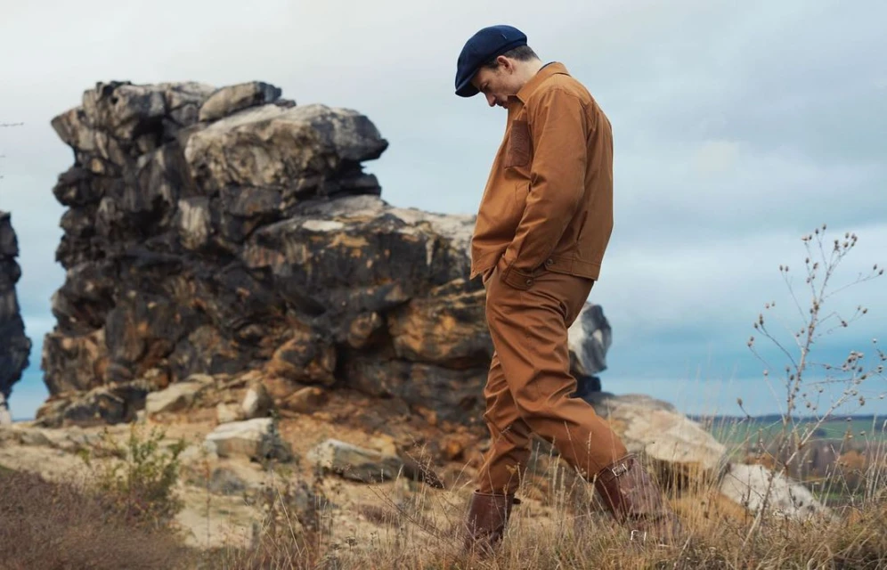 Adult male wearing brown jacket and pants walks in a rural outdoor setting with rock formations and dry vegetation under an overcast sky.