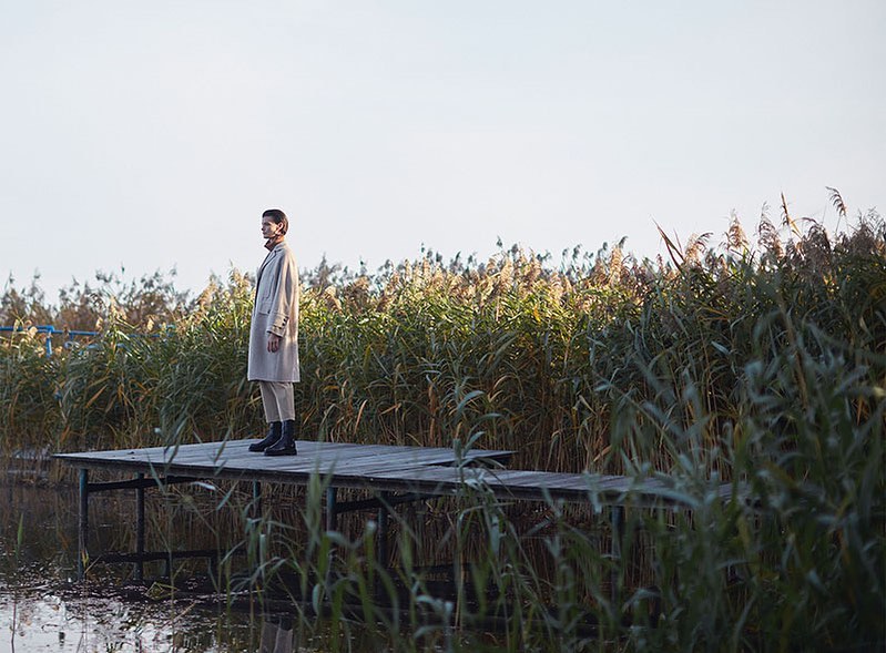 A fashionable adult woman stands on a weathered wood dock, wearing a coat, pants, and boots, with tall grass and a bright sky.