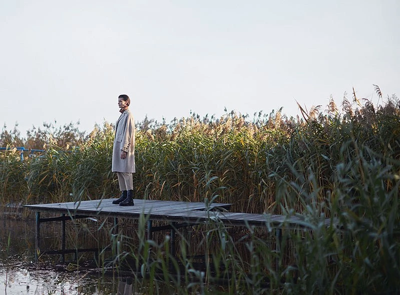 A fashionable adult woman stands on a weathered wood dock, wearing a coat, pants, and boots, with tall grass and a bright sky.