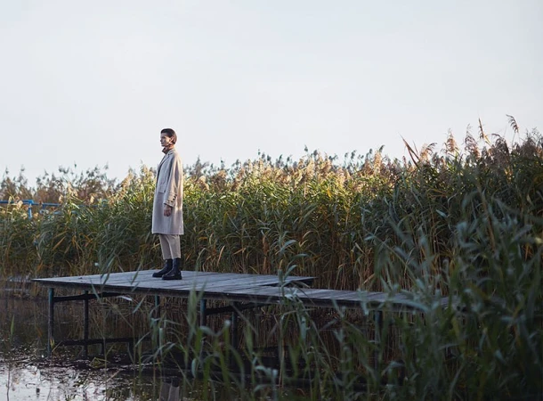 A fashionable adult woman stands on a weathered wood dock, wearing a coat, pants, and boots, with tall grass and a bright sky.
