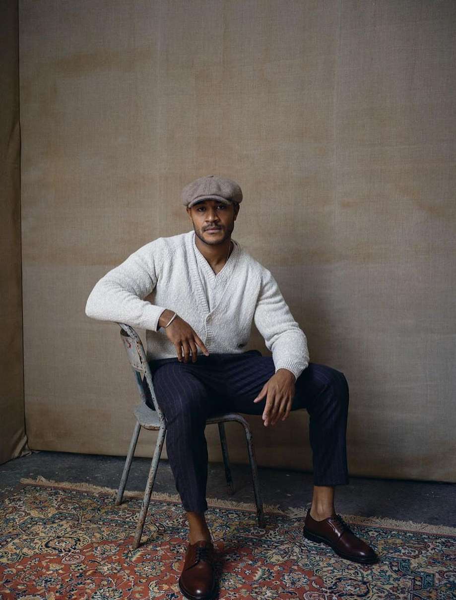 A man sits in a vintage-style chair on a patterned rug in a neutral setting wearing a cap, knit cardigan, striped trousers, and leather shoes.