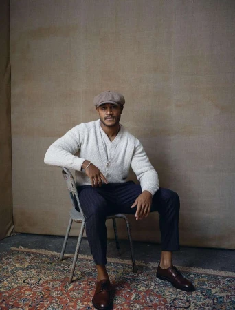 A man sits in a vintage-style chair on a patterned rug in a neutral setting wearing a cap, knit cardigan, striped trousers, and leather shoes.
