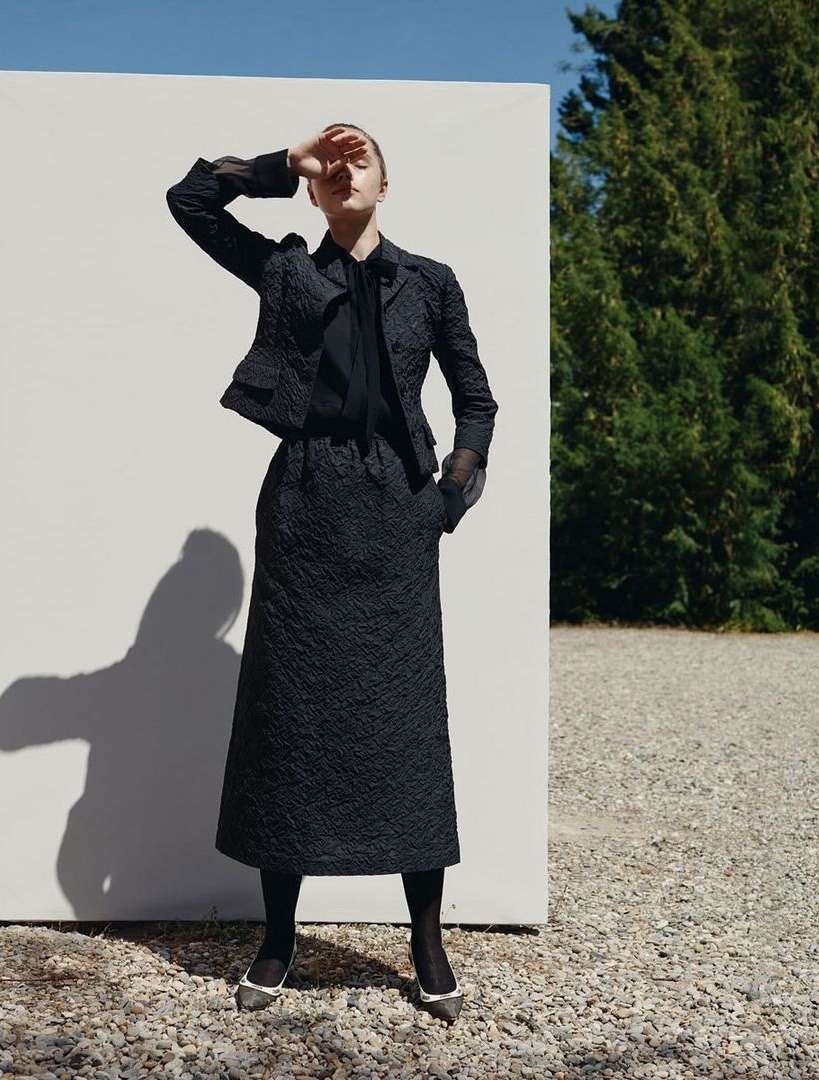 A woman in a dark textured skirt suit poses outdoors with a white wall, trees, and gravel in the background, shielding her eyes.