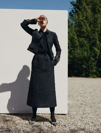 A woman in a dark textured skirt suit poses outdoors with a white wall, trees, and gravel in the background, shielding her eyes.