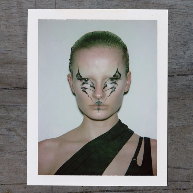 Striking portrait of a model with graphic black-and-white makeup on a white background, wearing a draped black garment, studio lit.