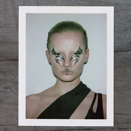 Striking portrait of a model with graphic black-and-white makeup on a white background, wearing a draped black garment, studio lit.