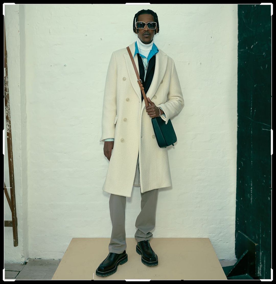Full body shot of an adult male posing on a platform in a white overcoat, grey pants and black shoes with a bag.