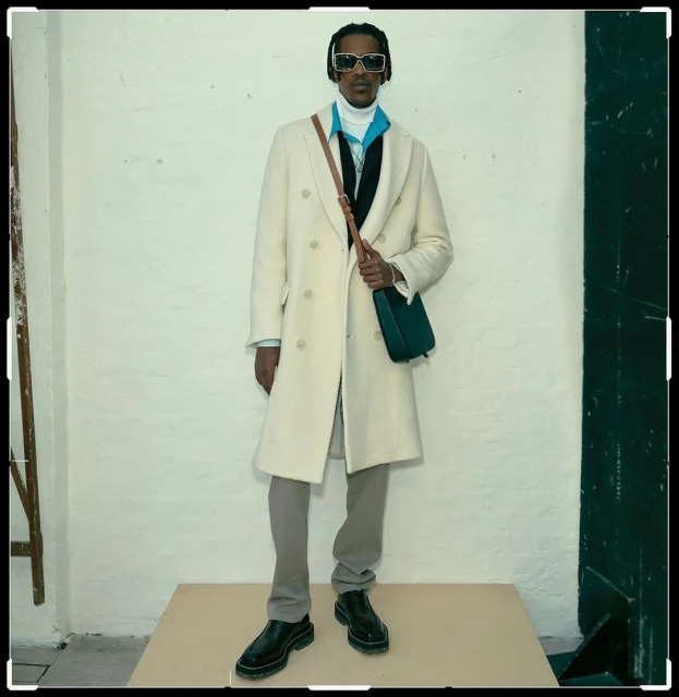 Full body shot of an adult male posing on a platform in a white overcoat, grey pants and black shoes with a bag.