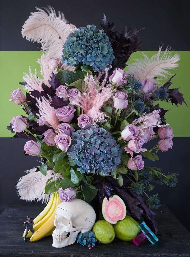 Still life featuring a vibrant floral arrangement with hydrangeas and roses, alongside a skull, bananas, and guavas on a dark table.