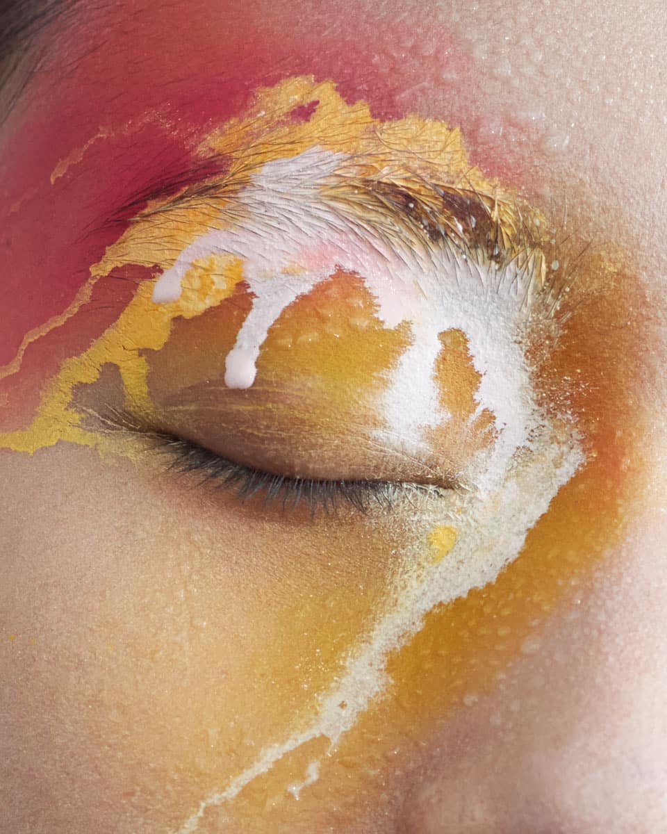 Extreme close-up of a face with artistic makeup in red, yellow, and white. The makeup covers the eyelid and cheek.