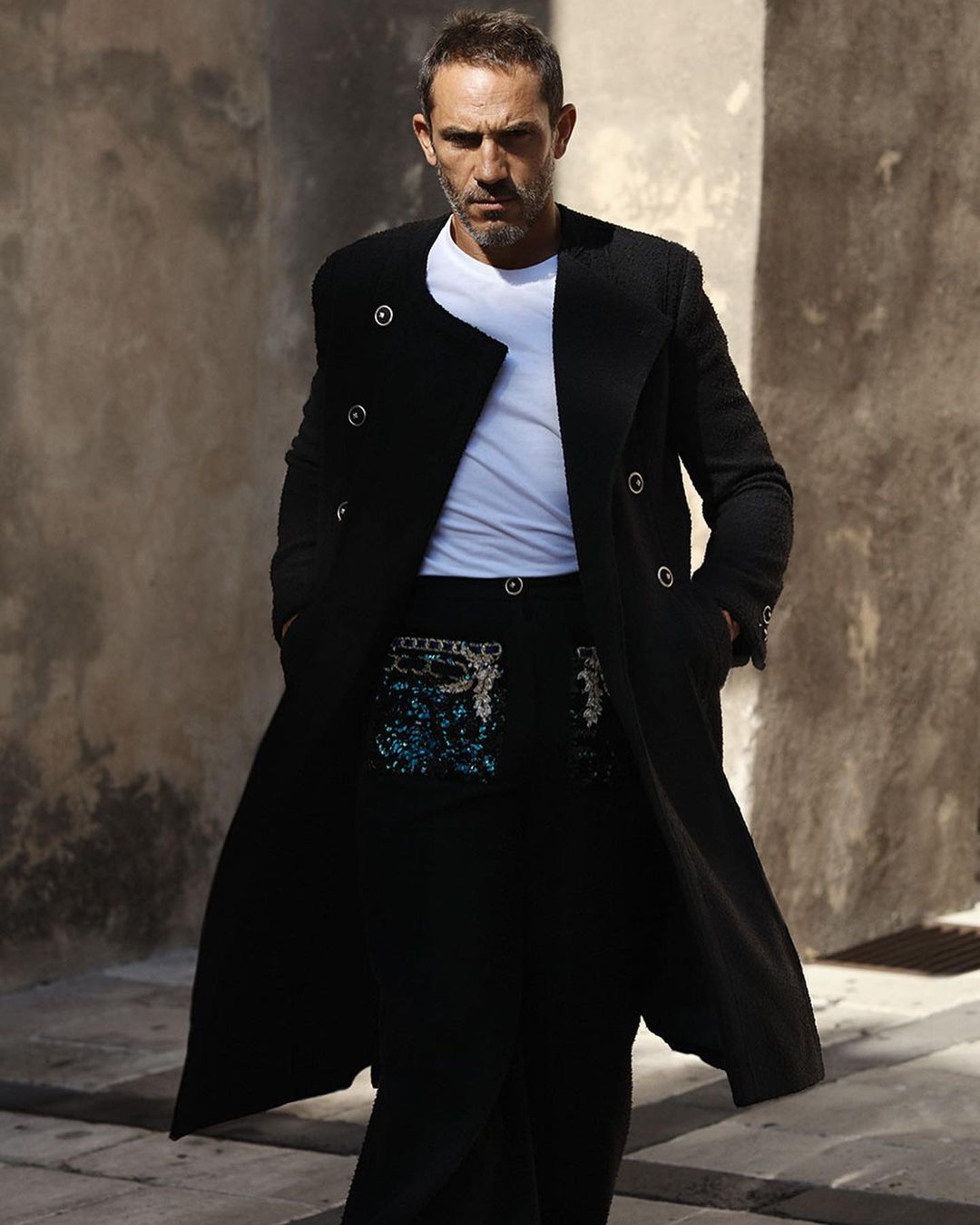 Stylish adult male in a long black coat and white t-shirt stands in front of a textured wall, posing in an urban setting.