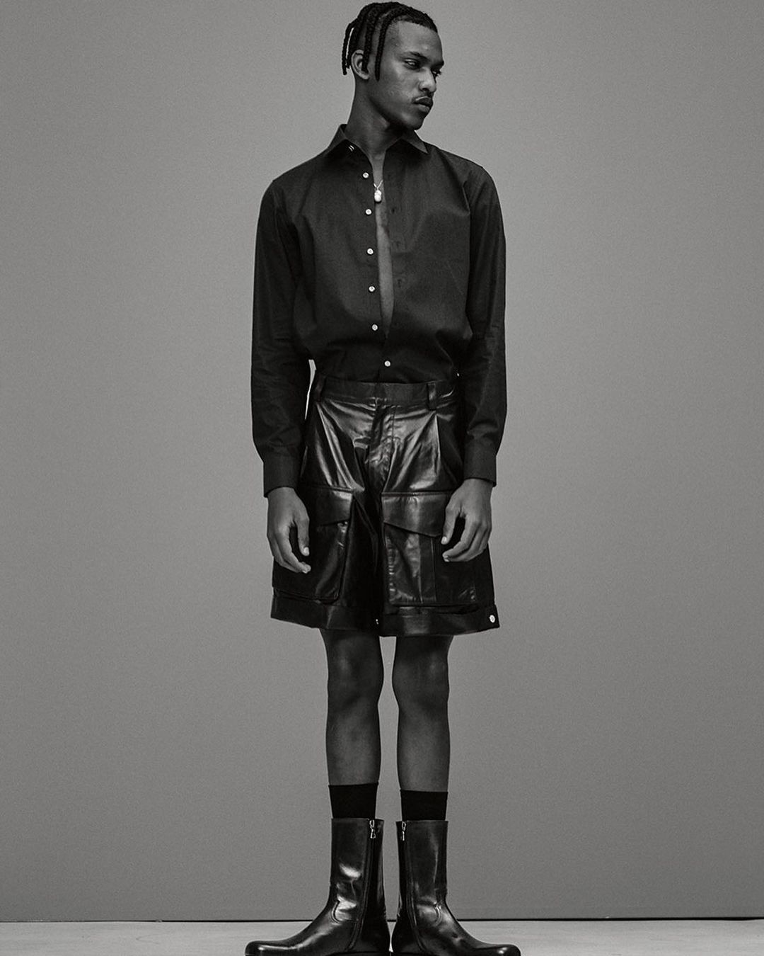 Portrait of a black male model in a black unbuttoned shirt and black leather shorts and boots, posing in a full shot against a grey background.