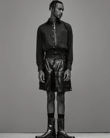 Portrait of a black male model in a black unbuttoned shirt and black leather shorts and boots, posing in a full shot against a grey background.