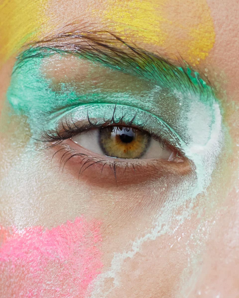 Close-up portrait of an adult female's eye with green, yellow, and pink makeup around the eye and face, creating an artistic and colorful look.