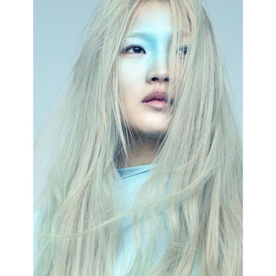 Portrait of an adult female with fair skin and long white hair posing indoors with her face painted blue in an eye-level shot.