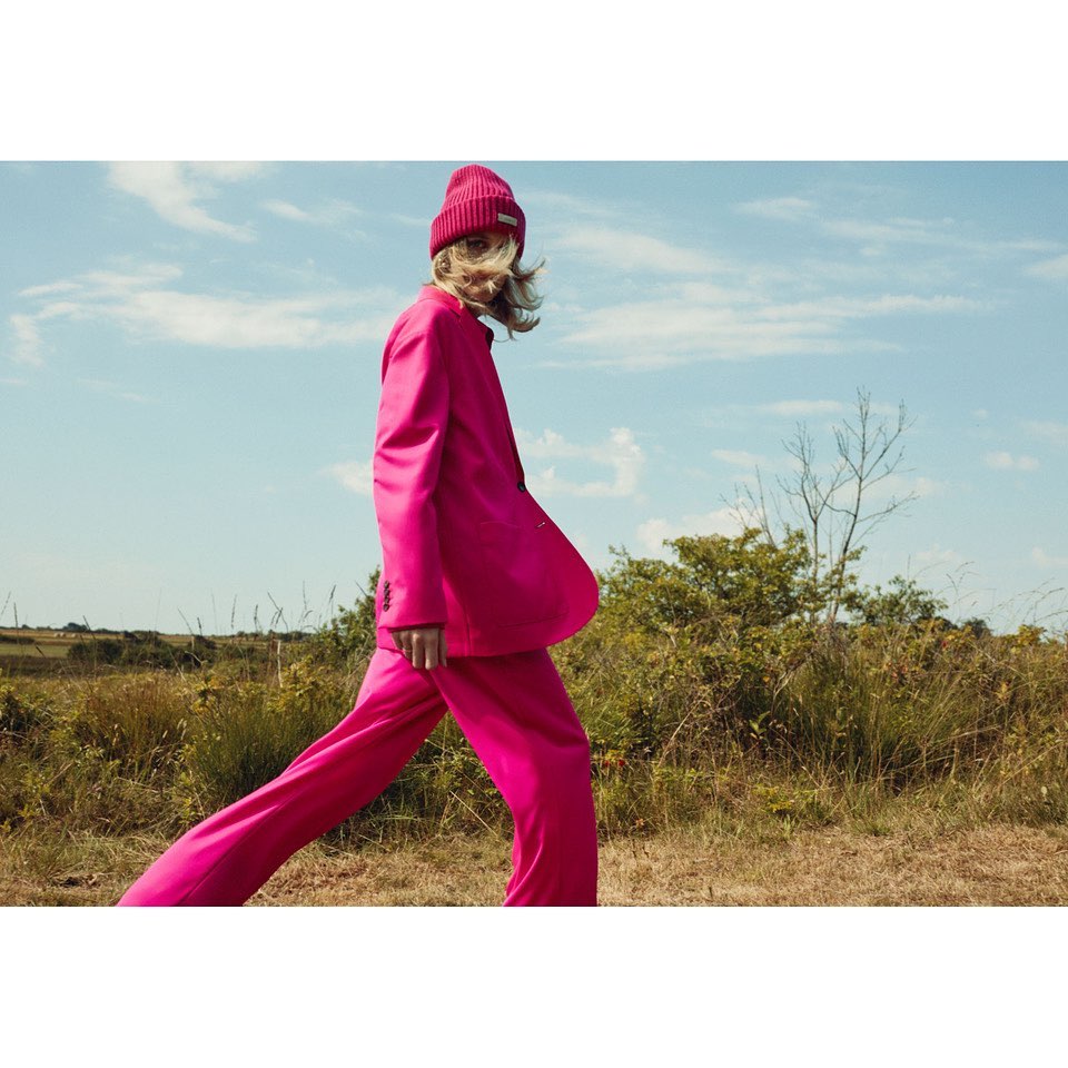 A stylish adult woman walks across a grassy field in a pink suit and matching beanie hat, set against a clear blue sky.
