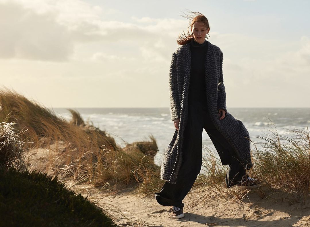 A woman in a long coat stands on a sandy dune near the ocean, with blowing grass and soft sunlight.
