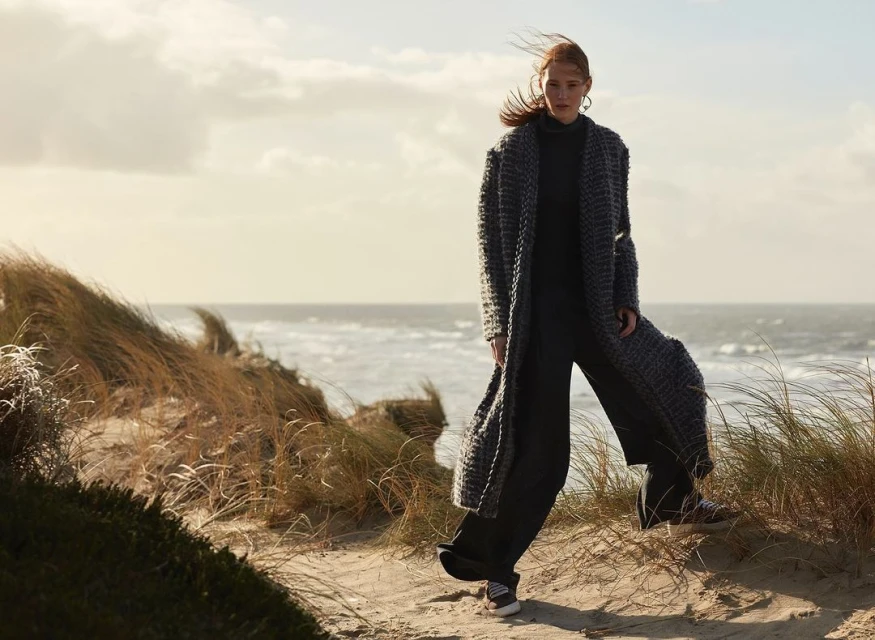 A woman in a long coat stands on a sandy dune near the ocean, with blowing grass and soft sunlight.