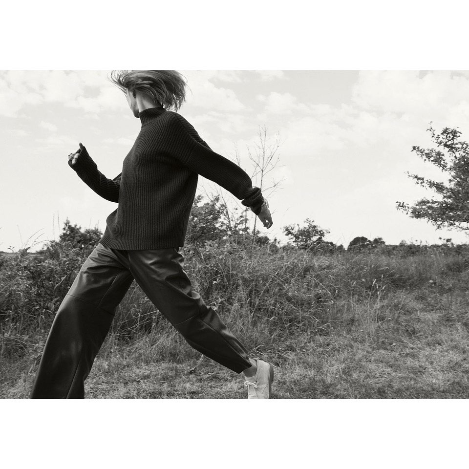 Monochrome image of a woman in motion wearing a dark sweater and pants outdoors in a grassy field under a cloudy sky.