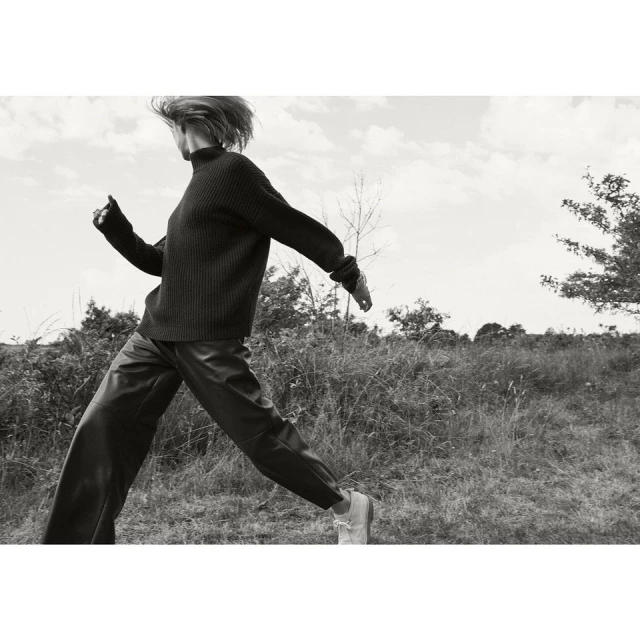 Monochrome image of a woman in motion wearing a dark sweater and pants outdoors in a grassy field under a cloudy sky.