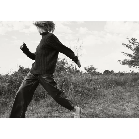 Monochrome image of a woman in motion wearing a dark sweater and pants outdoors in a grassy field under a cloudy sky.